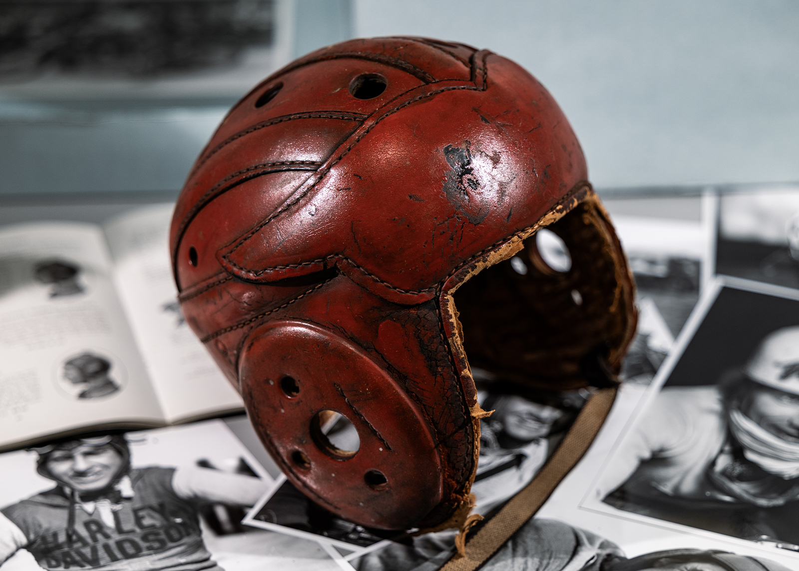 A vintage leather motorcycle helmet with a wing shape on the front is displayed on a table surrounded by black and white archival photos of riders.